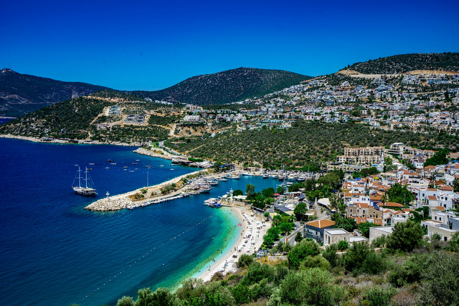 A small harbour with a stone breakwater and a sandy beach below white houses on green hills on the Antalya coast
